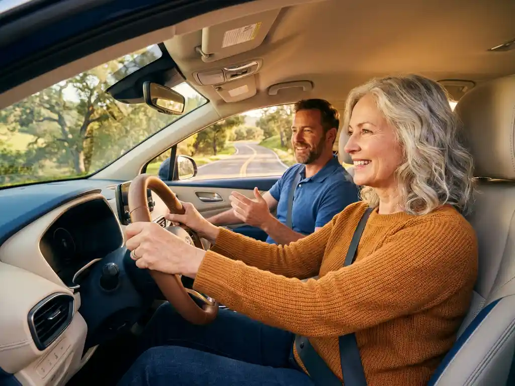 Älterer Erwachsener am Steuer eines modernen Autos lächelt einem Fahrlehrer zu, sonnige Landstraße im Hintergrund.