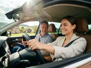 Junger Erwachsener lächelt beim Autofahren, Fahrlehrer auf Beifahrersitz, grüne deutsche Landschaft im Hintergrund.