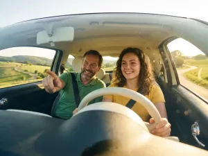 Lächelnde junge Fahrschülerin am Steuer eines modernen Autos bei sonniger Fahrstunde im Odenwald, Fahrlehrer auf dem Beifahrersitz.