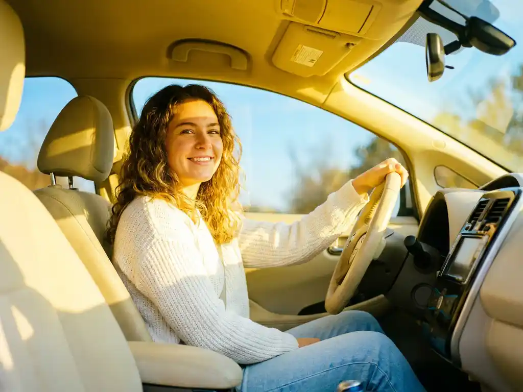 Junge Frau lächelt entspannt am Steuer eines modernen Autos im warmen Morgenlicht.