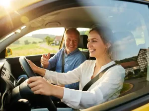 Junge Frau am Steuer lächelt zuversichtlich, Fahrlehrer sitzt daneben, goldenes Sonnenlicht durchflutet das Fahrzeuginnere, grüne Landschaft im Hintergrund.