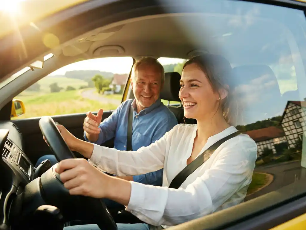 Junge Frau am Steuer lächelt zuversichtlich, Fahrlehrer sitzt daneben, goldenes Sonnenlicht durchflutet das Fahrzeuginnere, grüne Landschaft im Hintergrund.