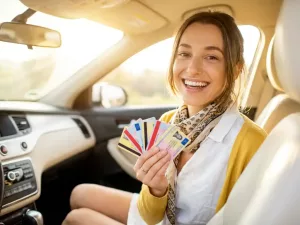 Lächelnde junge Frau hält mehrere bunte Führerscheinkarten im Auto, warmes Sonnenlicht durch die Windschutzscheibe.