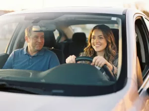 Junge Frau lächelt entspannt am Steuer eines modernen Autos, Fahrlehrer nickt aufmunternd, goldenes Sonnenlicht durchflutet die Windschutzscheibe.