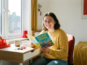 Junger Erwachsener studiert lächelnd ein buntes Theorieheft am hellen Schreibtisch, Autoschlüssel liegen daneben, Sonnenlicht fällt durchs Fenster.
